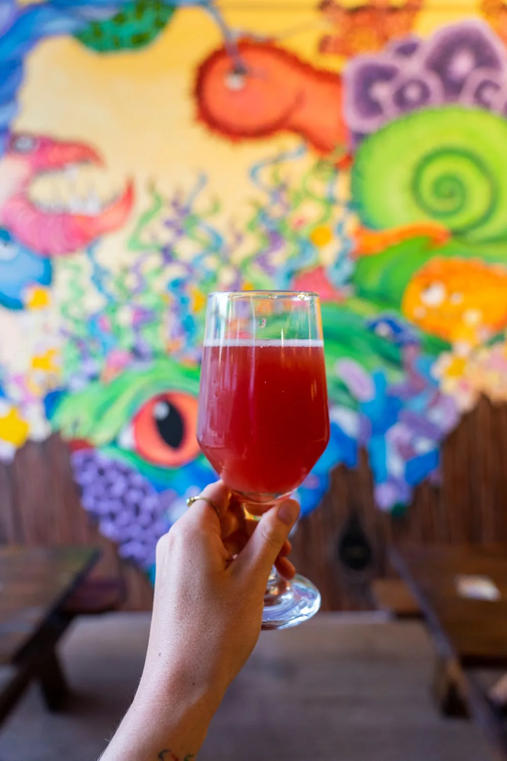 Woman holding red-colored sour beer in front of art mural inside Little Animals Brewery in downtown Johnson City TN