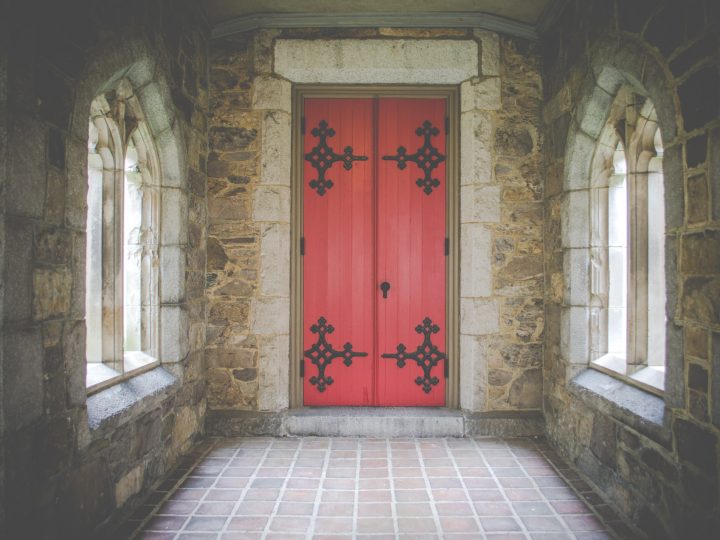 Stone walls and triangular windows with red ornate door inside church.