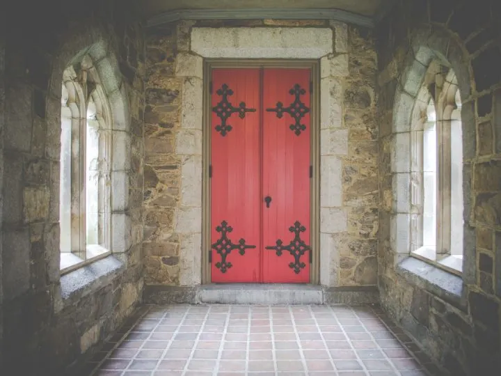 Stone walls and triangular windows with red ornate door inside church.