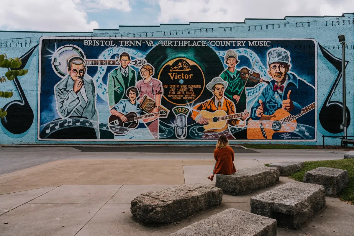 Woman sitting on rock bench in front of blue painted mural of the Birthplace of Country Music in historic downtown Bristol Tennessee-Virginia.