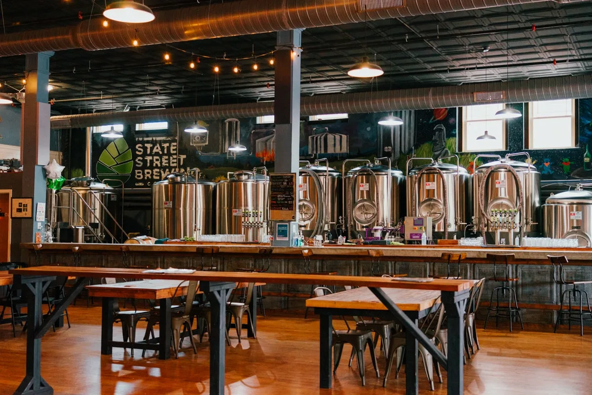 Inside the State Street Brewery with stainless steel beer barrels and wooden tables.