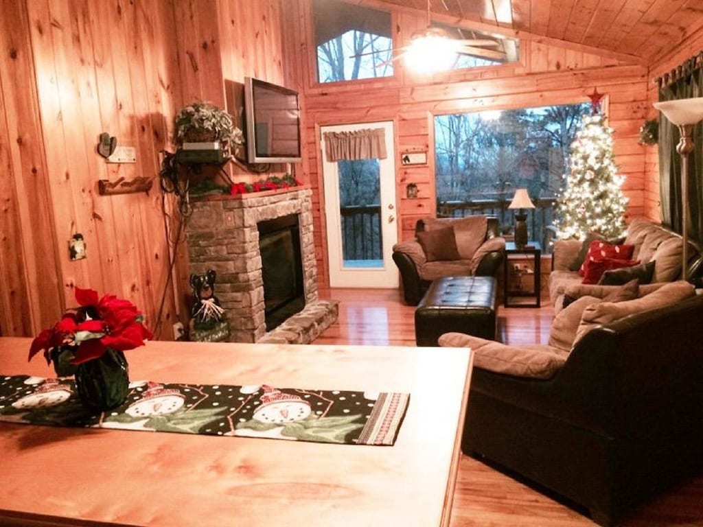 living room of a cozy cabin rental with large screen tv above a fireplace and a lit christmas tree. 