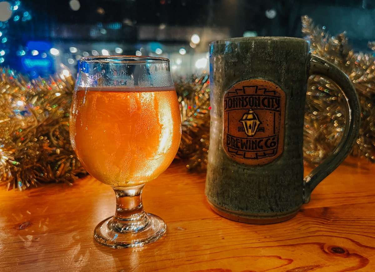 Half-pour of craft beer and mug of Johnson City Brewing Co with Christmas decorations in window.