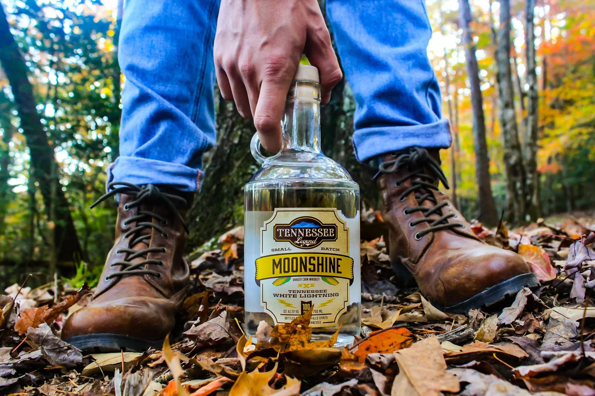 man holding a bottle of white lightning moonshine from tennessee legend distillery on the ground with fall foliage